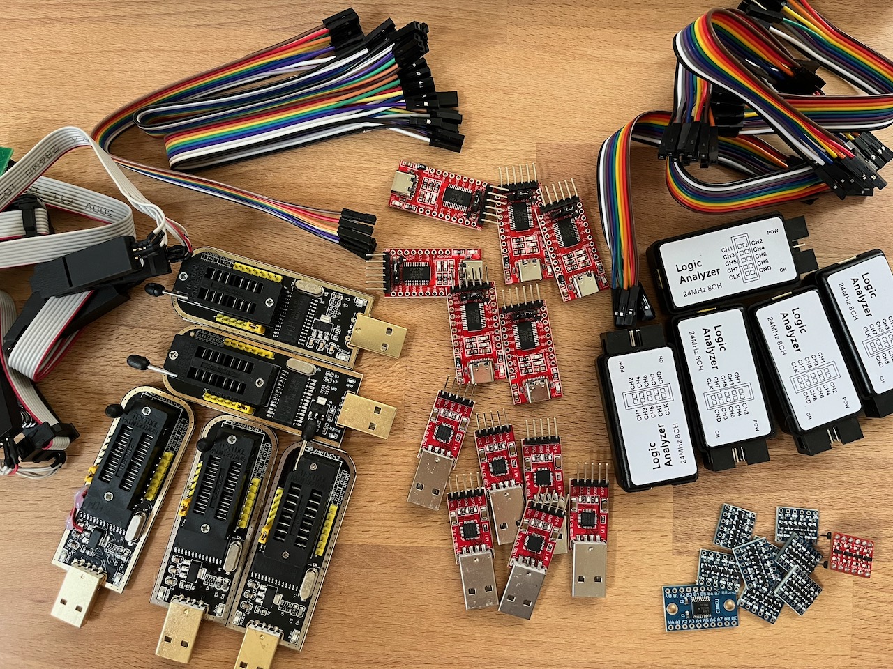 The hardware hacking kit spread on a table: CH341A flash programmers with SOIC-8 clips, CP2102 USB-UART adapters, 24 MHz 8-channel logic analyzers, jumper wires, and breakout boards.