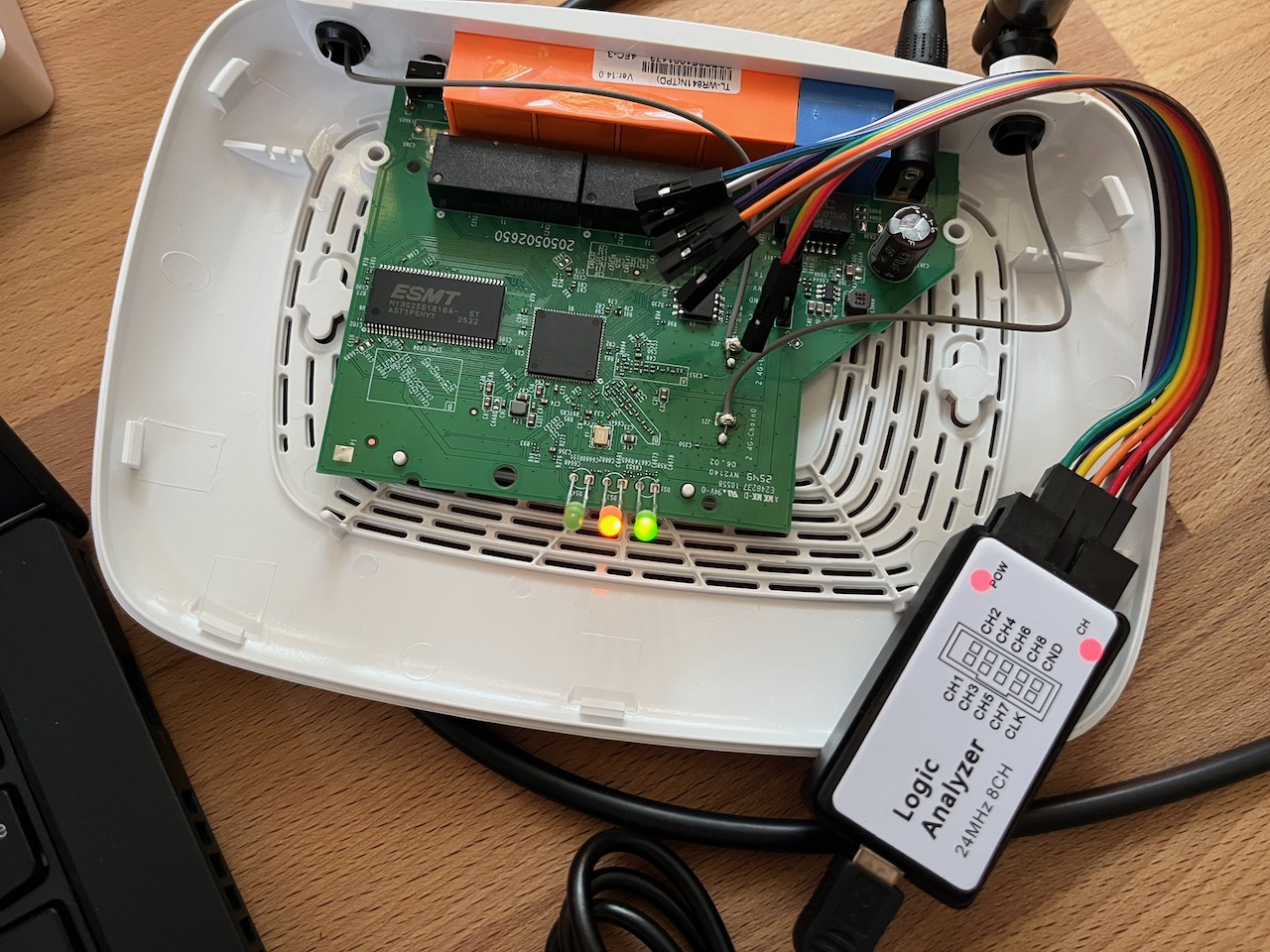 A TP-Link TL-WR841N opened on the bench, its PCB exposed, with a logic analyzer clipped to the UART header via jumper wires and the status LEDs lit.